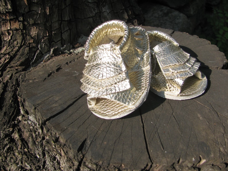May include: A pair of gold sandals with a ruffled design. The sandals are made of a textured material and have a strap around the ankle.
