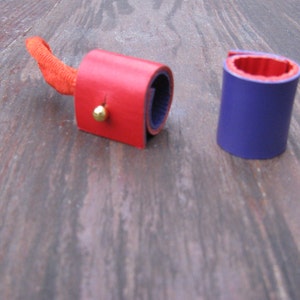 May include: Two leather wrist cuffs, one red and one purple. The red cuff has a gold button and a small orange strap. The purple cuff has a red interior.