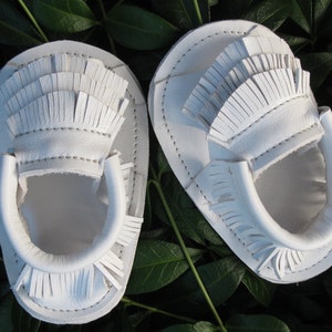 May include: A pair of white leather baby moccasins with fringe detailing. The moccasins are made of soft leather and have a comfortable fit.