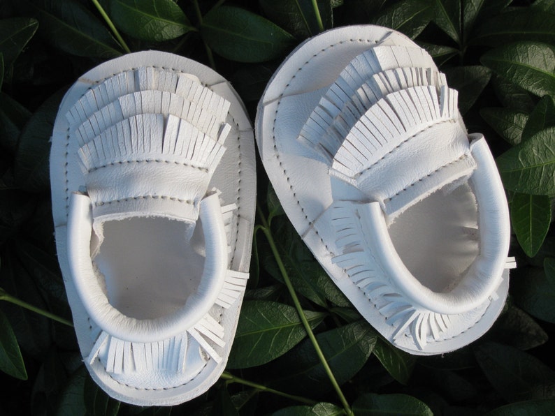May include: A pair of white leather baby moccasins with fringe detailing. The moccasins are made of soft leather and have a comfortable fit.