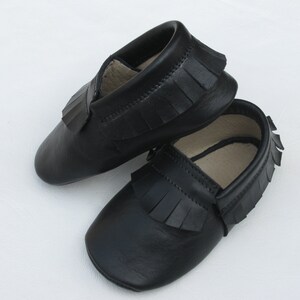 Puede incluir: Un par de mocasines de beb&eacute; de cuero negro con flecos. Los zapatos son de suela blanda y tienen un dise&ntilde;o cl&aacute;sico, perfectos para beb&eacute;s. Los mocasines se muestran sobre un fondo blanco.