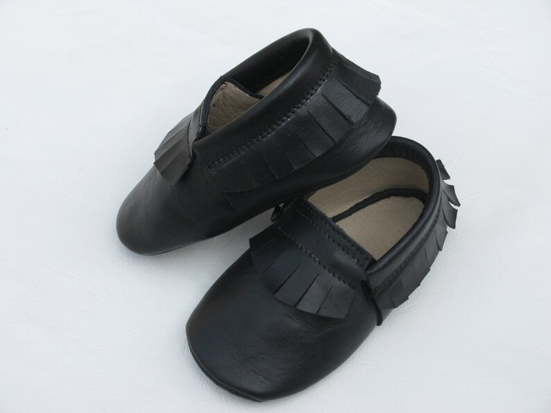 Puede incluir: Un par de mocasines de beb&eacute; de cuero negro con flecos. Los zapatos son de suela blanda y tienen un dise&ntilde;o cl&aacute;sico, perfectos para beb&eacute;s. Los mocasines se muestran sobre un fondo blanco.
