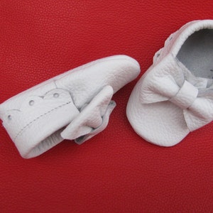 May include: Two white leather baby shoes with a bow on the front. The shoes are on a red background.
