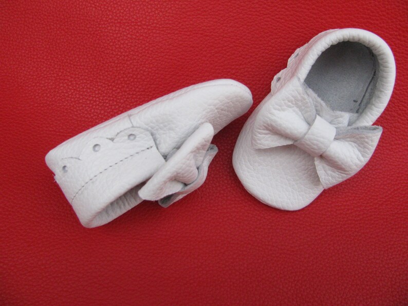 May include: Two white leather baby shoes with a bow on the front. The shoes are on a red background.