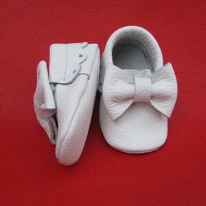 May include: A pair of white leather baby shoes with a bow on the front. The shoes are on a red background.
