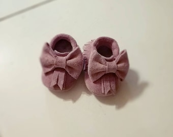 May include: A pair of soft, mauve-colored baby moccasins. Each shoe features a large bow on the front and a fringed detail. The shoes are made of a suede-like material, perfect for a newborn.