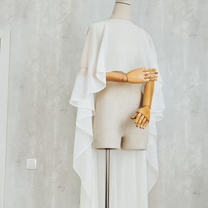 May include: A white sheer cape with a high-low hemline draped over a beige mannequin. The garment features a wide, flowing silhouette with layered fabric panels that extend down the back, creating a soft, translucent appearance on the torso.