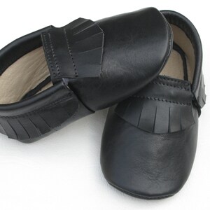 Puede incluir: Un par de mocasines de beb&eacute; de cuero negro. Los zapatos tienen una suela blanda y un detalle de flecos en la parte superior. Los mocasines est&aacute;n dise&ntilde;ados con un estilo sin cordones y probablemente sean para beb&eacute;s o ni&ntilde;os peque&ntilde;os.