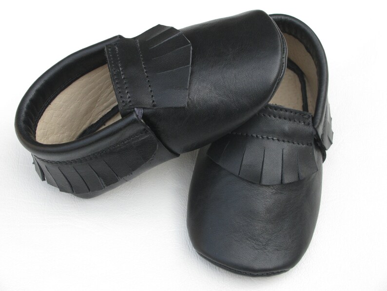Puede incluir: Un par de mocasines de beb&eacute; de cuero negro. Los zapatos tienen una suela blanda y un detalle de flecos en la parte superior. Los mocasines est&aacute;n dise&ntilde;ados con un estilo sin cordones y probablemente sean para beb&eacute;s o ni&ntilde;os peque&ntilde;os.