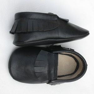 Puede incluir: Un par de mocasines negros de cuero para beb&eacute;. Cada zapato presenta una pala con flecos y un interior suave de color claro. Los zapatos est&aacute;n dise&ntilde;ados con una puntera redondeada y un estilo sencillo y cl&aacute;sico.