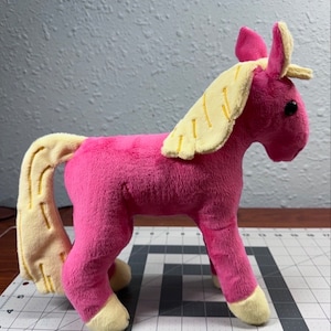 Puede incluir: Juguete de caballo de peluche rosa con una melena y cola amarillas. El caballo está de pie sobre una superficie de rejilla gris y blanca.