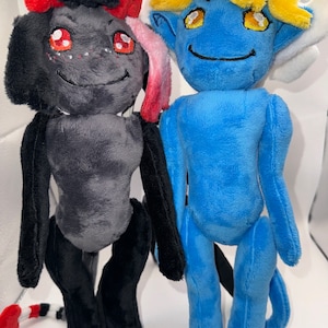 Op de afbeelding: Twee pluche speelgoedjes, een zwart met rode hoorns en een staart, de andere blauw met geel haar en hoorns. Beide hebben geborduurde ogen en een glimlach.