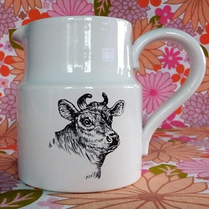 May include: White ceramic pitcher with a black and white illustration of a cow's head. The pitcher has a curved handle and a spout for pouring. The background is a floral pattern.