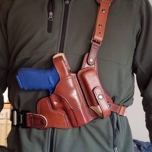May include: A brown leather shoulder holster with a blue handgun inside. The holster is worn over a green jacket and secured with a black belt.