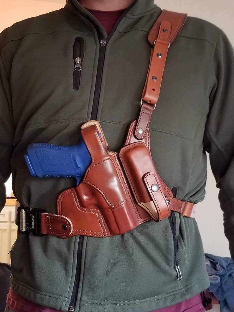 May include: A brown leather shoulder holster with a blue handgun inside. The holster is worn over a green jacket and secured with a black belt.