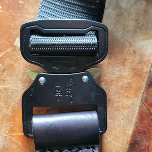 May include: Black and silver adjustable buckle with a black nylon strap and a black leather strap with a stitched edge. The buckle has a brass slider and the text "COBRA" is printed on the buckle.