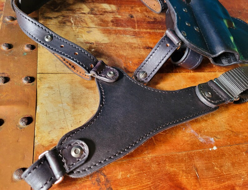 May include: A black leather holster with adjustable straps for carrying a firearm. The holster has a smooth, polished finish and is designed for concealed carry.