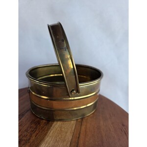 May include: A small, oval-shaped brass bucket featuring a polished finish and a central horizontal copper band. The container includes a single, upright, hinged handle attached to the sides with small metal rivets, showcasing a metallic sheen.