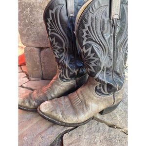 Peut inclure: Une paire de bottes de cowboy en cuir noir et gris avec des motifs de surpiqûres grises complexes sur les tiges hautes, des bouts arrondis, des talons bottiers et des coutures apparentes le long des semelles des chaussures.