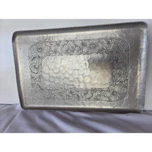 May include: A rectangular silver-toned metal tray featuring a hammered texture on the central surface and a raised border decorated with an embossed floral pattern. The tray has rounded corners and a smooth, metallic finish throughout the design.