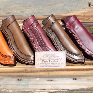 May include: Five leather knife sheaths in different colors, including brown, orange, and burgundy. The sheaths are decorated with stitching and have a pointed tip. A business card for "DnA Leather" is in the center of the image.