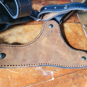 May include: A brown leather shoulder holster with black stitching and metal rivets. The holster is designed to carry a handgun.