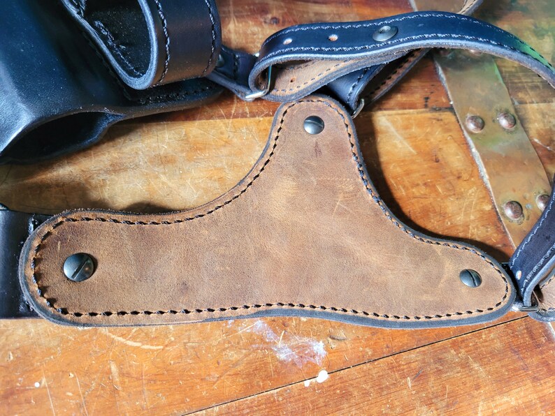 May include: A brown leather shoulder holster with black stitching and metal rivets. The holster is designed to carry a handgun.
