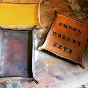 May include: Two leather trays, one brown and one tan. The tan tray has the words "PHONE WALLET KEYS" embossed on it.