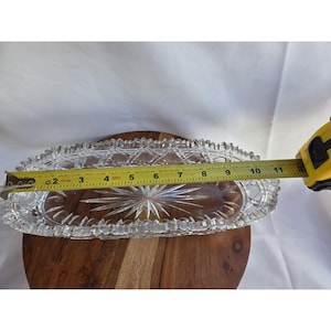 May include: An oval clear glass dish featuring a faceted diamond pattern along the sides and a starburst design at the center. A yellow measuring tape is positioned across the dish, showing a length of approximately 11.5 inches.