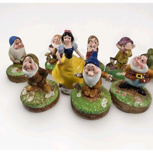 Puede incluir: Una colección de figuras de cerámica que presenta a un personaje femenino central con un vestido azul y amarillo rodeado por siete figuras de enanos distintas, cada una sobre una base de hierba verde con flores blancas y bordes marrones.