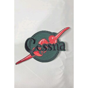 May include: A circular embroidered patch featuring a dark green background with a black border. The word Cessna is stitched in black lettering across the center, overlaid on a stylized red graphic element that extends diagonally across the circle.