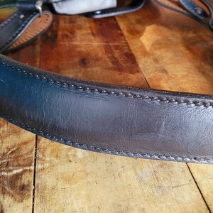 May include: A close-up of a black leather camera strap with stitching. The strap is curved and has a smooth surface.