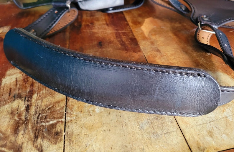May include: A close-up of a black leather camera strap with stitching. The strap is curved and has a smooth surface.