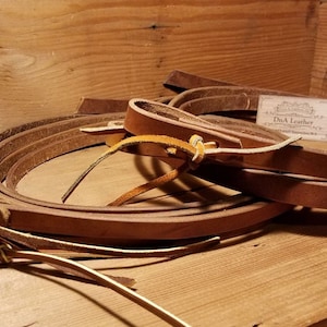 May include: A set of three brown leather reins with a D.A. Leather business card. The reins are folded and stacked on top of each other. The reins have a knotted loop at one end and a stitched leather tab at the other end.