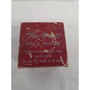 Op de afbeelding: Een vierkante rode doos voor een Tru-Point potloodslijper. De doos bevat zilverkleurige tekst: Tru-Point PENCIL POINTER, Produces long, slender, true needle point; for fine line work of all kinds. Het rode oppervlak vertoont tekenen van slijtage.