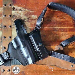 May include: A black leather shoulder holster with a magazine pouch. The holster is designed to carry a handgun and has a strap that goes over the shoulder.