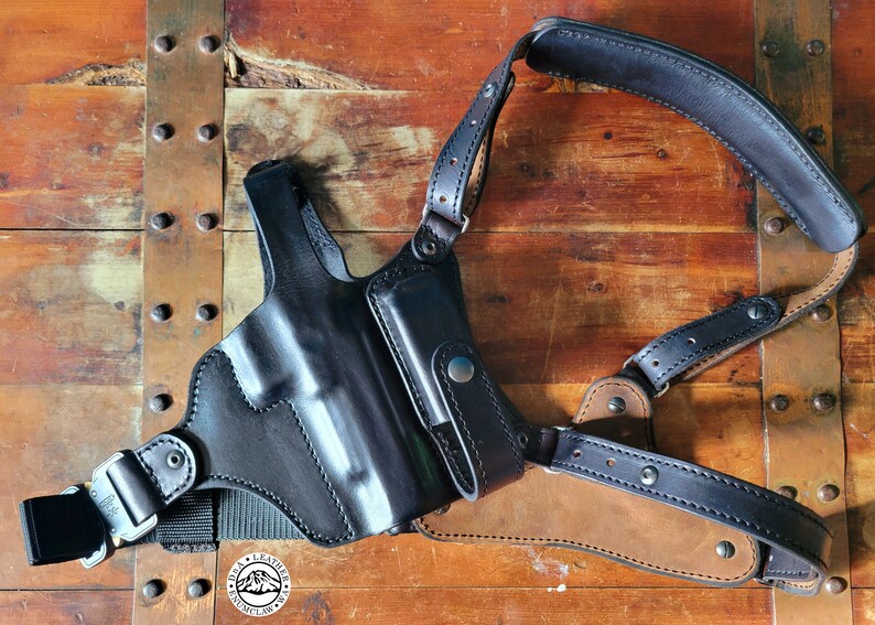 May include: A black leather shoulder holster with a magazine pouch. The holster is designed to carry a handgun and has a strap that goes over the shoulder.