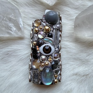 May include: A rectangular lighter decorated with an assortment of mixed media elements including a central realistic brown eye cabochon, various iridescent and metallic beads, clear faceted crystals, and small silver-toned metal charms.