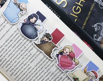May include: A collection of colorful, illustrated character bookmarks, featuring various figures and objects. The bookmarks are placed on the pages of a book, with some sticking out. The bookmarks have a cartoon style.