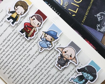 May include: A collection of colorful, illustrated character bookmarks. Each bookmark features a different cartoon character with unique outfits and accessories. The bookmarks are placed on the pages of a book.