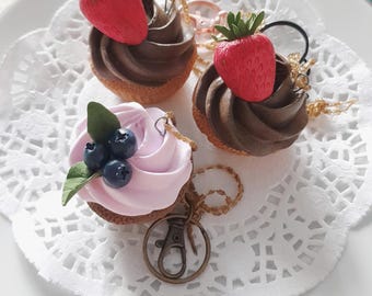 May include: Three miniature cupcake keychains with different toppings. One cupcake is topped with chocolate frosting and a red strawberry, another with chocolate frosting and a red strawberry, and the last with pink frosting and blueberries.