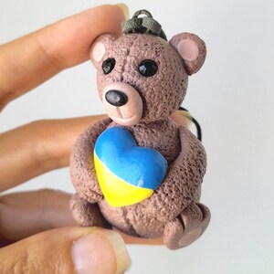 Può includere: Piccola statuetta di orsetto marrone che tiene un emblema a forma di cuore. Il cuore è dipinto di blu e giallo, i colori della bandiera ucraina. L'orso ha occhi neri e un piccolo naso.