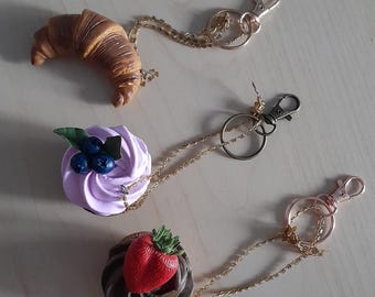 May include: Three keychains with different food charms: a croissant, a purple cupcake with blueberries, and a chocolate cupcake with a strawberry. Each charm is attached to a gold chain with a metal clasp.