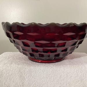 May include: A ruby red glass bowl with a diamond pattern and a scalloped rim. The bowl sits on a white towel. The bowl is a decorative piece.