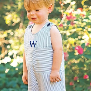 May include: A young child wearing a blue and white striped overall with a blue letter 'W' embroidered on the front. The child is standing in a garden with green foliage and pink flowers.