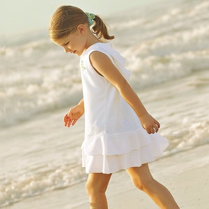 May include: A young girl in a white dress walks on a sandy beach with the ocean waves in the background.