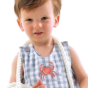 Könnte beinhalten: Ein kleines Kind trägt einen blauen und weißen karierten Latzhose mit einem roten Krebs, der auf der Vorderseite gestickt ist. Das Kind hält ein weißes Seil.