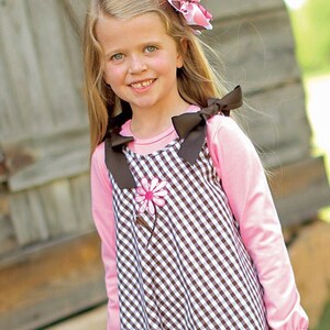 Peut inclure: Une jeune fille portant une robe en vichy marron et blanc avec une applique de fleur rose. La robe a un ourlet festonné et est portée sur une chemise rose à manches longues. La fille porte des collants marrons et a un nœud marron dans les cheveux.