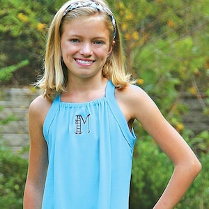 Puede incluir: Una persona joven lleva un vestido azul claro sin mangas con un dobladillo con volantes. El vestido tiene una sola inicial, "M", bordada en la parte delantera.