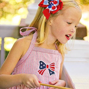 May include: A young girl wearing a red and white striped dress with a red, white and blue bow in her hair. The dress has a red, white and blue bow with stars on it.